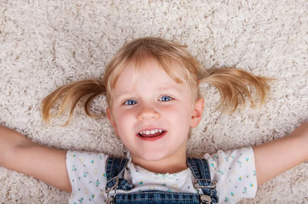 Sonriente linda niña acostada en la alfombra blanca y mirando camer — Foto de Stock