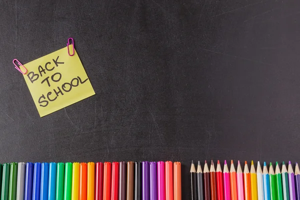 Повернутися до шкільного фону з барвистими фломастерами, олівцями та заголовком "Назад до школи", написаними на жовтому аркуші паперу на шкільній дошці — стокове фото