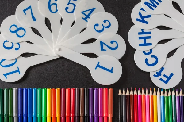 Back to school background with a lot of colorful felt-tip pens and colorful pencils and cards of numerals from one to ten and consonant letters of alphabet on the  chalkboard