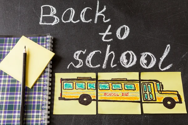 Назад до школи з назвою "Назад до школи", написаною крейдою на чорній дошці та зображенням шкільного автобуса, намальованого на жовтих шматочках паперу та блокнота з олівцем — стокове фото