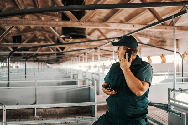 勤勤恳恳的农民穿着工作服 用智能手机给动物经销商打电话 谷仓里没有动物 — 图库照片