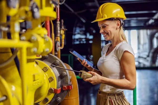 Smiling hardworking female supervisor in working uniform with helmet on head standing holding tablet and checking on heat on pipe while standing in heating plant. Woman doing male job concept.