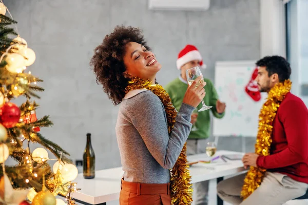 クリスマスツリーの横に立って シャンパンを保持し 彼女の会社でクリスマスの前夜に素晴らしい時間を持っている混合レースの実業家の笑顔 背景には彼女の同僚が祝う — ストック写真