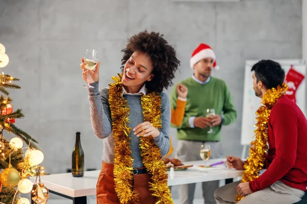 クリスマスツリーの横に立って シャンパンを保持し 彼女の会社でクリスマスの前夜に素晴らしい時間を持っている混合レースの実業家の笑顔 背景には彼女の同僚が祝う — ストック写真