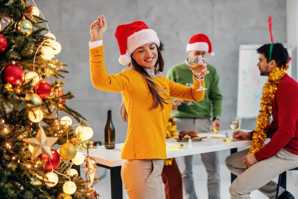 サンタの帽子をかぶった陽気な若い実業家は 彼女の会社でクリスマスツリーの横にシャンパンダンスを保持しています クリスマスを祝う彼女の同僚もいる — ストック写真