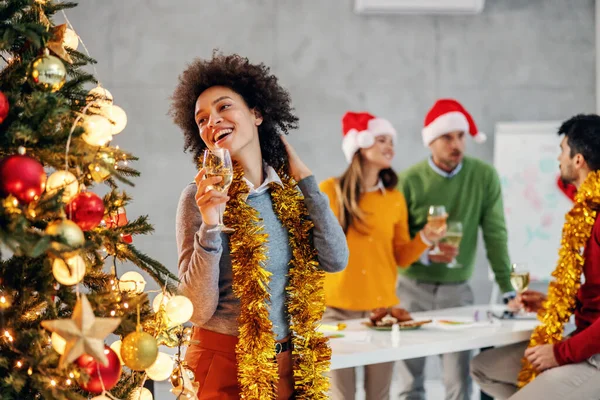クリスマスツリーの横に立って シャンパンを保持し 彼女の会社でクリスマスの前夜に素晴らしい時間を持っている混合レースの実業家の笑顔 背景には彼女の同僚が祝う — ストック写真
