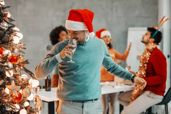 クリスマスツリーの隣でサンタの帽子をかぶった若いビジネスマンとクリスマス前夜に彼の会社でシャンパンを飲む 背景には 彼の同僚も祝う — ストック写真