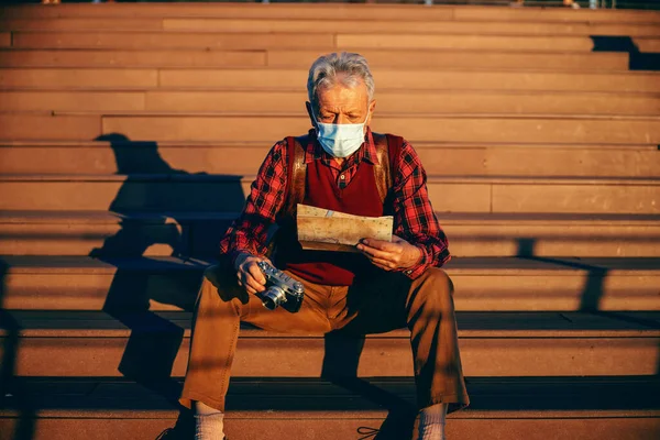 ファッショナブルなシニア男性の上に保護面を着用し 階段に座っている 男はカメラを保持し 都市マップを見て コロナウイルスの概念中の観光 — ストック写真