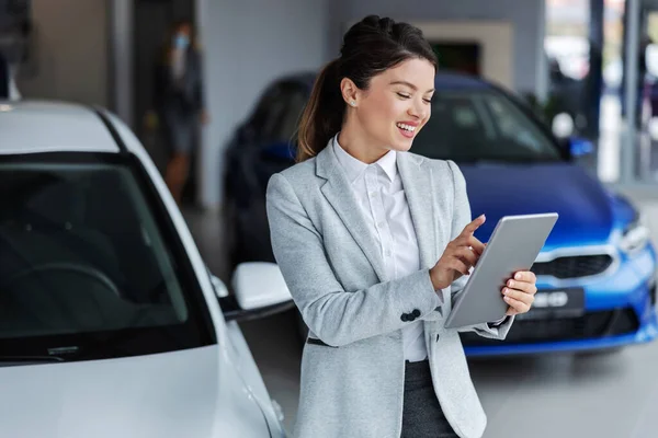 Smiling female seller in suit using tablet for looking which car is sold while standing in car salon.
