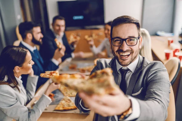テーブルに座ってピザのスライスを保持眼鏡に合わせてハンサムな笑顔の白人ビジネスマン 背景に彼の同僚を食べることです 昼休み — ストック写真