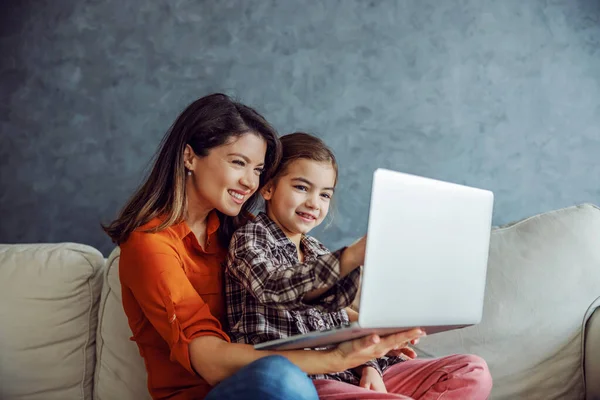 Happy mother and daughter sitting on sofa and using laptop. Mother holding daughter in her lap while daughter pointing at laptop.