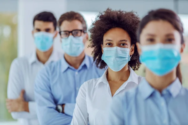 Multicultural group of business people with face masks on standing in office with arms crossed and looking at camera. Selective focus on mixed race woman in the middle..