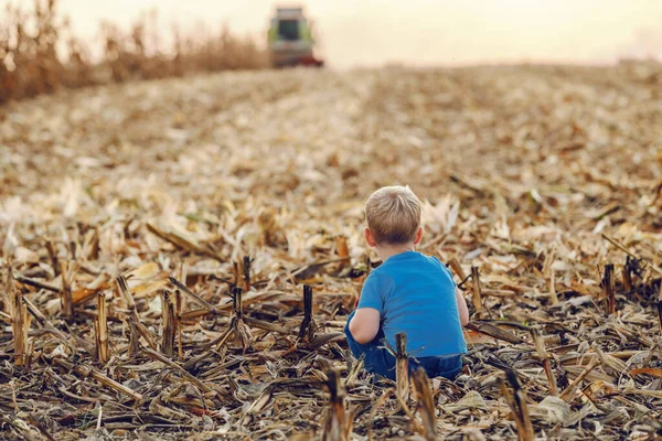 一个可爱的农民小男孩蹲在玉米地上玩耍的后视镜 背景是收割机收获 — 图库照片