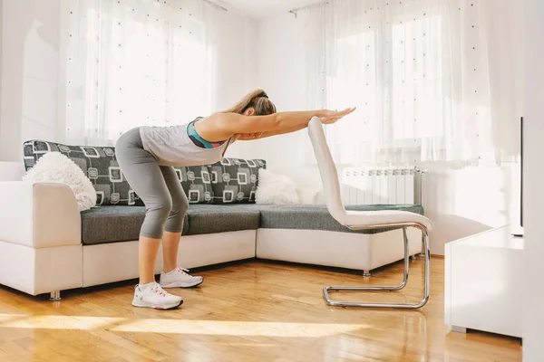 Muscular sportswoman in shape doing fitness exercises for her backs while leaning on chair. If you can\'t go to the gym, you can make your own at home.