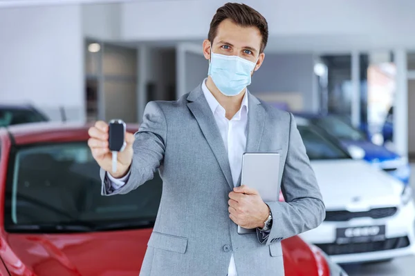 Car seller with face mask standing in car salon and showing keys on a new brand car which are ready to be sold.