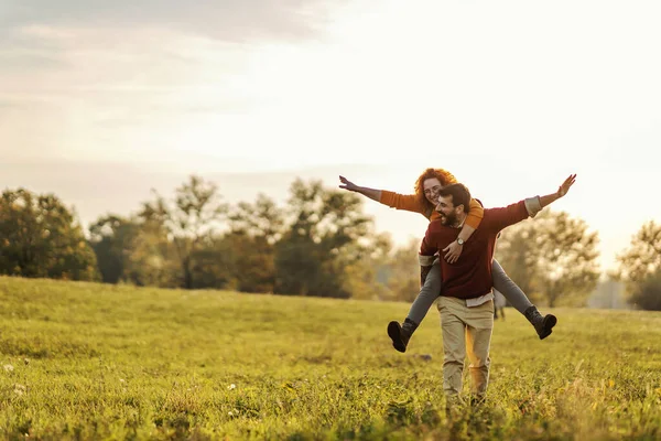 Young happy playful couple in love having piggyback ride. Man holding woman on his backs and walking on a meadow on a beautiful autumn sunny day.