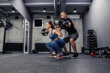 Personal training in the gym. A young woman and in sportswear and in good shape, she does barbell squats to strengthen the muscles of the whole body. The assistance of trainers in individual training clipart