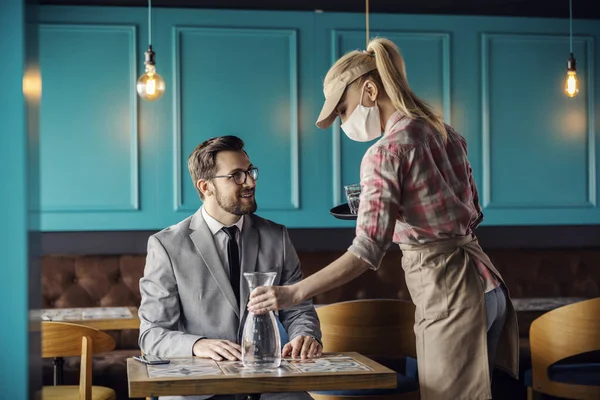 Restaurant service and corona virus. The blonde waitress is wearing a uniform and a protective face mask. She puts a bottle of water on the table and serves the guest in a suit in a modern restaurant