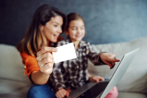 Mother and daughter sitting on the sofa and using laptop for online shopping. Mother is holding credit card while daughter pointing at toy she wants. Selective focus on credit card.