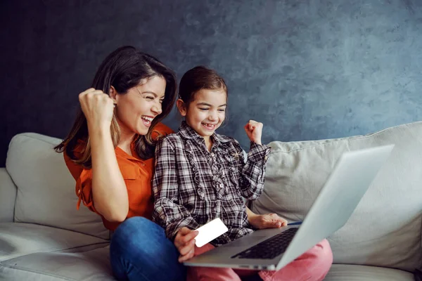 Smiling mother and daughter sitting on sofa and using laptop for online shopping. Daughter holding credit card. They had successful purchase so they are overjoyed and holding fists.