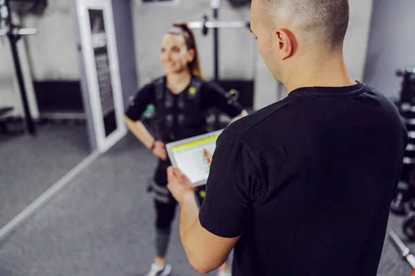 トレーニングセンターの真ん中に立つ黒のEmsトレーニングスーツ姿の女と トレーニングパフォーマンスのあるデジタルタブレットを持つトレーナーの背中の一部 現代のコーチングコンセプト — ストック写真