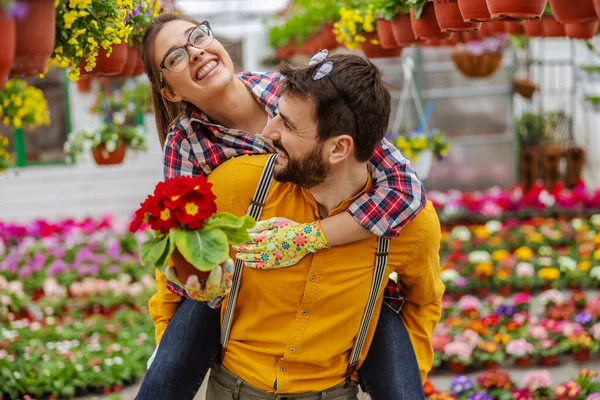 温室でピギーバックに乗る愛の笑みを浮かべてカップル 周りには色とりどりの花が咲いています 花を持つ女 小規模事業主 — ストック写真