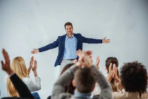 The motivation of employees in training. Smiling motivational speaker standing in front of his audience who is clapping. A multicultural group of businesspeople cheering and applauding a life coach