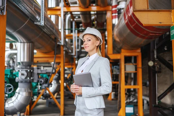 A professional female director in formal attire, with a protective helmet on her head holding a tablet and standing in a heating plant. She stands at the entrance of the heating plant. CEO female