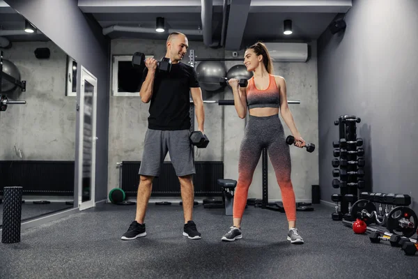 Fitness couple doing arm exercises together and lifting dumbbells indoor modern gym. Front view of a couple dressed in sportswear doing arm exercises and they make eye contact to support each other