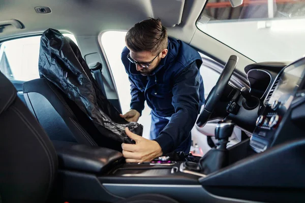 A worker in a car tries to put the upholstery on a car seat. Dressed in a blue work suit, he puts upholstery in the car. Man at work cleans the interior of a car. Car preparation, technical inspection