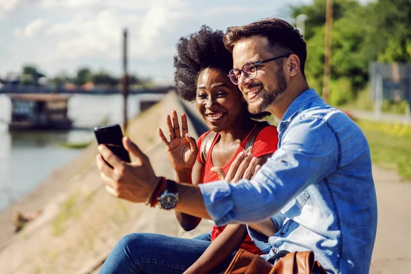 かわいい多文化のカップルが川のそばに座って スマートフォンで友人とビデオ通話をして — ストック写真