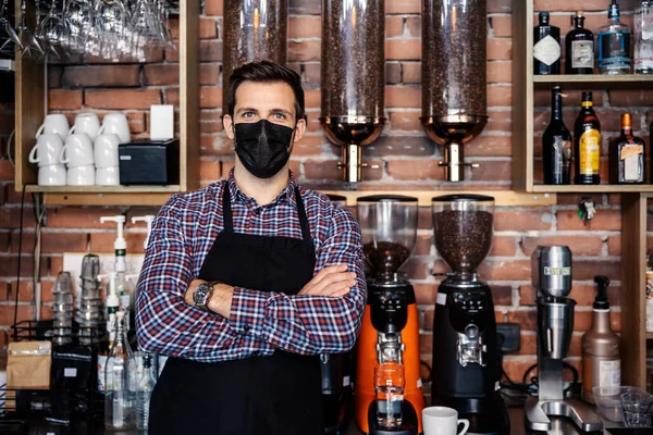 Drink service during the corona virus pandemic. The man stands at the restaurant counter and wears a plaid shirt and apron. A waitress has her arms crossed a wearing face protective mask