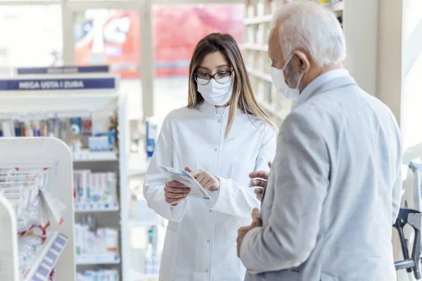 Help from a pharmacist. A female pharmacist with glasses holds medicine boxes and instructions for use. She looks at the man who is explaining something else. They both wear protective masks