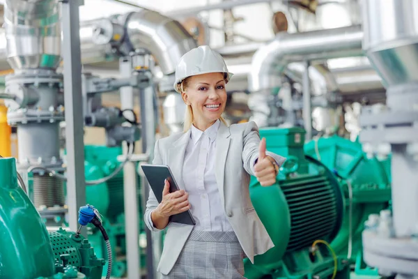Successfully completed inspection in the heating plant. Middle-aged blond female supervisor in suit with protective helmet on head standing in power plant with tablet in hands and showing thumbs up