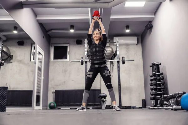 Confidence and powerful movement, body stability. Female in sportswear does exercise, lifting kettle bell weights in a special modern EMS suite in a modern gym concept. Electrical muscle stimulation