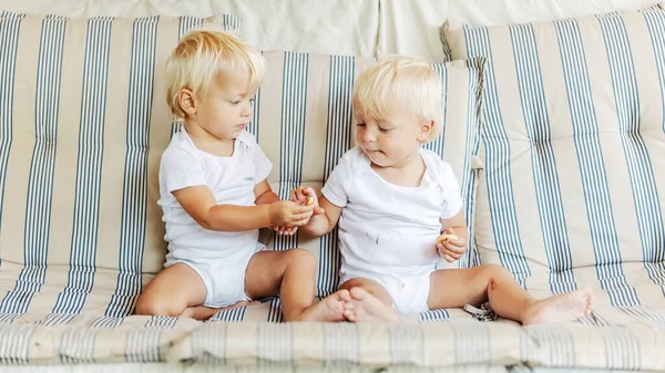 双胞胎宝宝共享一块饼干 两个穿着白色婴儿套装 沾满巧克力的孩子坐在客厅里舒适的米黄色沙发上 满含爱心的眼神 家庭幸福的时刻 — 图库照片