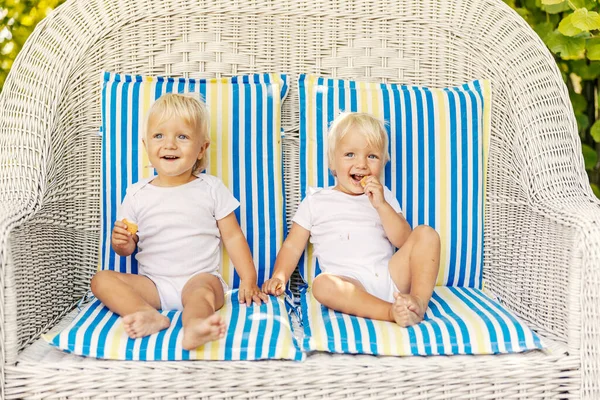 Sweet twins, a family full of love. The cute babies in the white bodysuits enjoy eating cookies and while sitting on a large canvas chair with colorful cushions. Twins with blue eyes and blonde hair