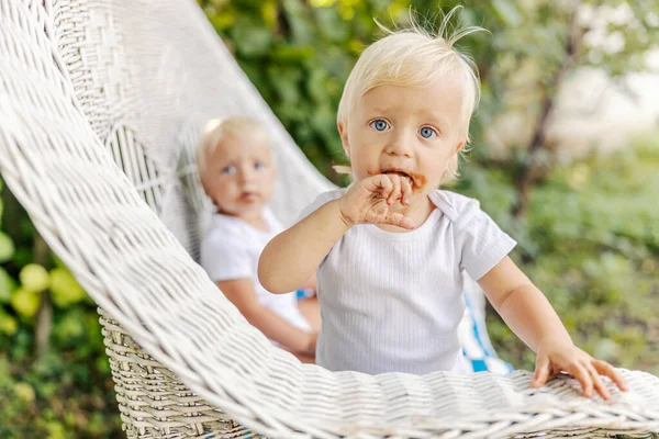 一个长着蓝眼睛和金发碧眼的小孩 长着一张肮脏的巧克力脸 坐在院子里的柳条椅上 看着摄像机 一个可爱的婴儿喜欢甜食 一个婴儿的滑稽照片 — 图库照片