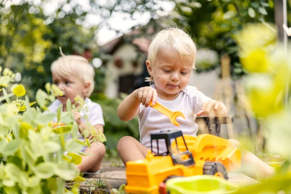 Playing in the garden with a plastic excavator game. Toddlers twins sit in the yard and play with plants. A child with blond hair and big blue eyes looks playing with their favorite toys