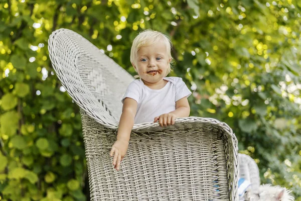 不器用な赤ん坊がチョコレートで微笑んだ 赤ちゃんは子供時代を楽しんでいます 大きなキャンバスベージュの椅子の側にもたれて白いボディスーツの青い目の子供 庭での遊びを楽しむ — ストック写真