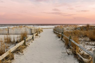 Akşamın ilerleyen saatlerinde, batan güneşten gelen pembe gökyüzü ile sahile giden bir patikanın fotoğrafı..