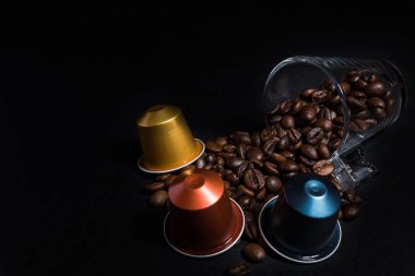 Kahve çekirdeği ve kahve fincanı ile ev yapımı kahve kapsülü makinelerinin soyut ve kavramsal hali. Siyah arka planda izole edilmiş espresso kahve kapsülleri koleksiyonu, yakın plan görüntü detayları. Kopyalama alanı olan grafiksel kaynaklar.