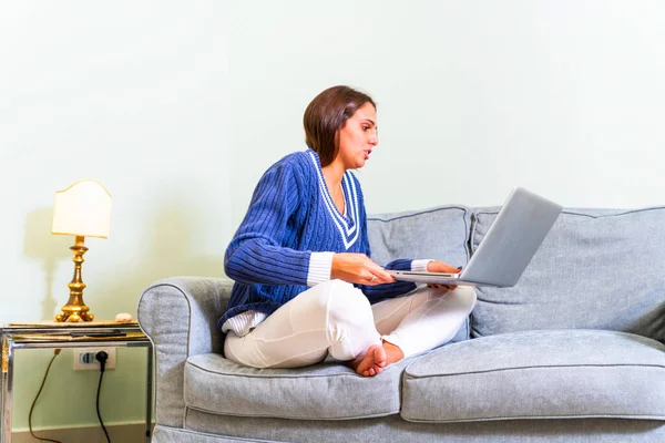 Happy young woman sitting on the sofa with crossed legs and using laptop on gray background. Image of cheerful american woman using laptop while sitting on sofa in living room.