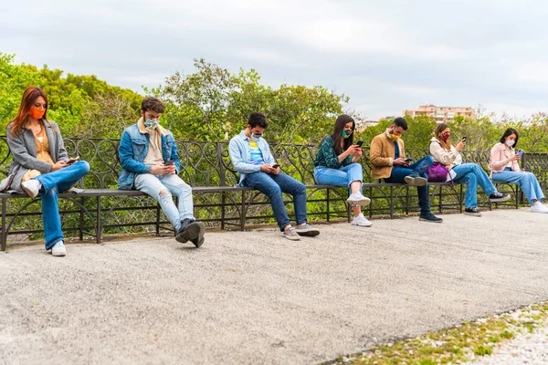 Młodzi Ludzie Siedzą Ławce Parku Noszenie Maski Twarz Używanie Smartfona — Zdjęcie stockowe