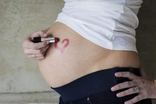 Mujer embarazada joven dibujando un corazón de lápiz labial en el vientre — Foto de Stock