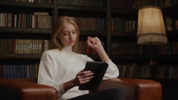 Молода жінка сидить у шкіряному кріслі та купує онлайн на планшеті з великою кількістю книг на фоні — стокове відео