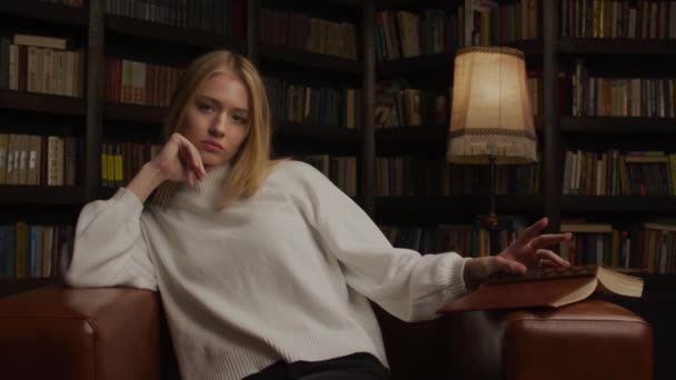 Μέτριο πλάνο ενός έφηβου ξανθού μοντέλου που ποζάρει και κοιτάζει κατευθείαν στην κάμερα σε ένα δωμάτιο γεμάτο βιβλία. — Αρχείο Βίντεο