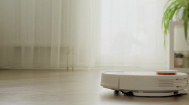 Beyaz bir elektrikli süpürge robotu parlak bir apartmanın içindeki ahşap zemini temizliyor.