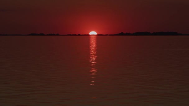 Solen sitter lågt över horisonten och reflekteras i en flod i en röd solnedgång — Stockvideo