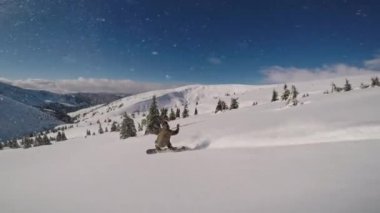 Bir snowboardcu dağlarda güneşli bir günde dönüşler yaparken kar kameraya sıkılıyor.
